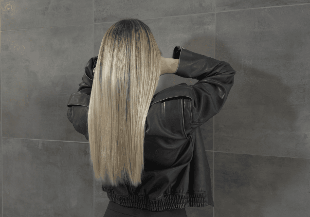 Soin Botox capillaire Jean Louis David sur cheveux longs et lisses, effet brillance intense et fibre capillaire réparée. Vue de dos d'une personne aux longs cheveux blonds parfaitement lisses et droits, tombant de manière uniforme sur le haut du dos. La chevelure blonde claire et lumineuse présente une texture soyeuse. La personne porte une veste en cuir noir structurée et se tient devant un mur en carrelage gris foncé.