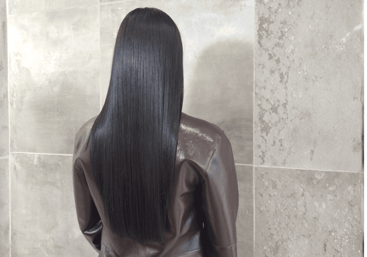 Soin Botox capillaire Jean Louis David sur cheveux longs et lisses, effet brillance intense et fibre capillaire réparée. Vue de dos d'une personne aux longs cheveux noirs parfaitement lisses et soyeux, tombant en pointe au milieu du dos. La chevelure présente une brillance intense et uniforme sous la lumière. Elle porte une veste en cuir marron et se tient devant un mur en carrelage beige.