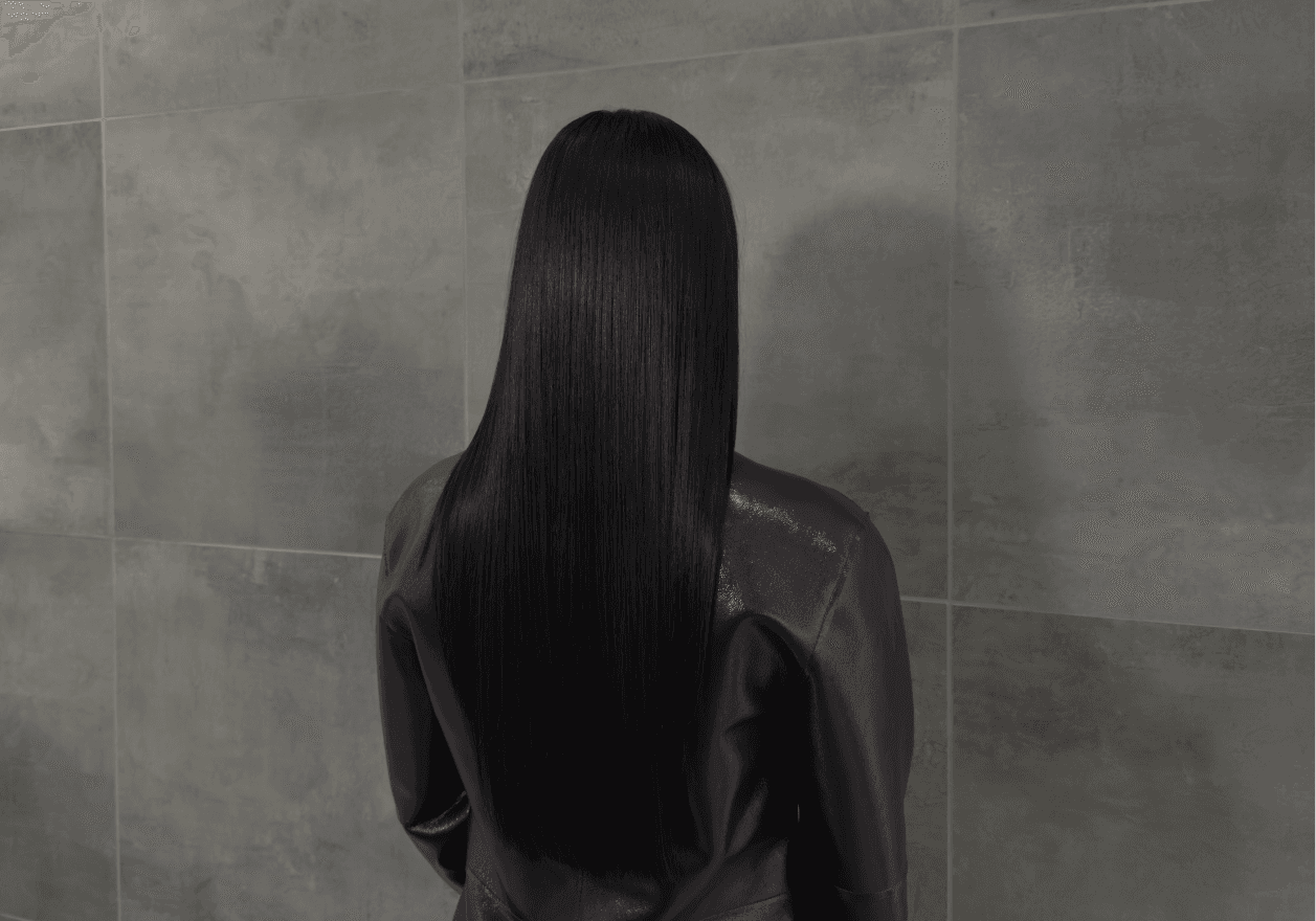 Vue de dos d'une personne aux longs cheveux noirs parfaitement lisses et soyeux, tombant en pointe au milieu du dos. La chevelure présente une brillance intense et uniforme sous la lumière. Elle porte une veste en cuir marron et se tient devant un mur en carrelage beige.