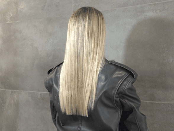 Vue de dos d'une personne aux longs cheveux blonds parfaitement lisses et droits, tombant de manière uniforme sur le haut du dos. La chevelure blonde claire et lumineuse présente une texture soyeuse. La personne porte une veste en cuir noir structurée et se tient devant un mur en carrelage gris foncé.