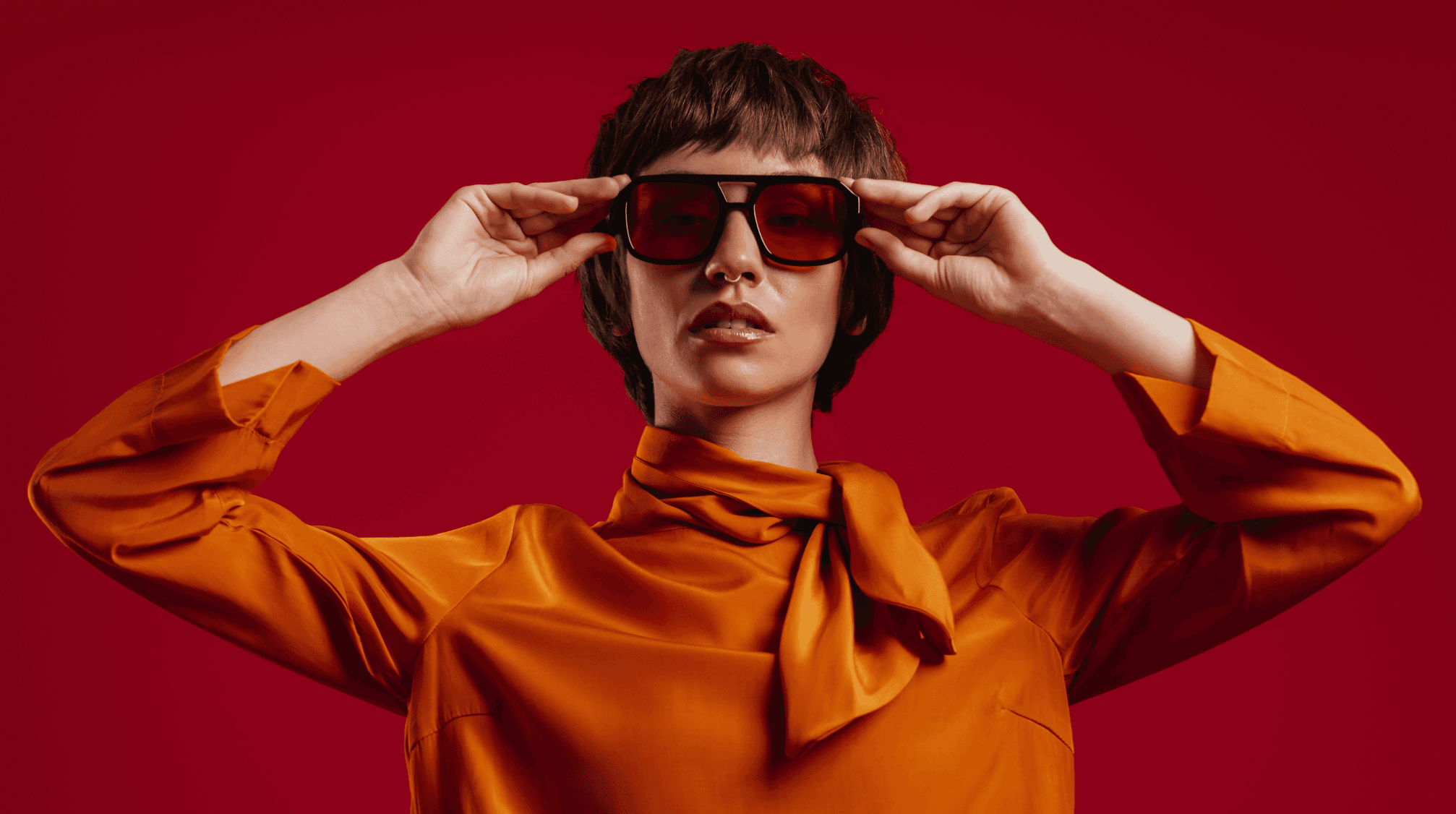 Portrait d'une jeune femme aux cheveux courts réglant ses lunettes sur fond rouge