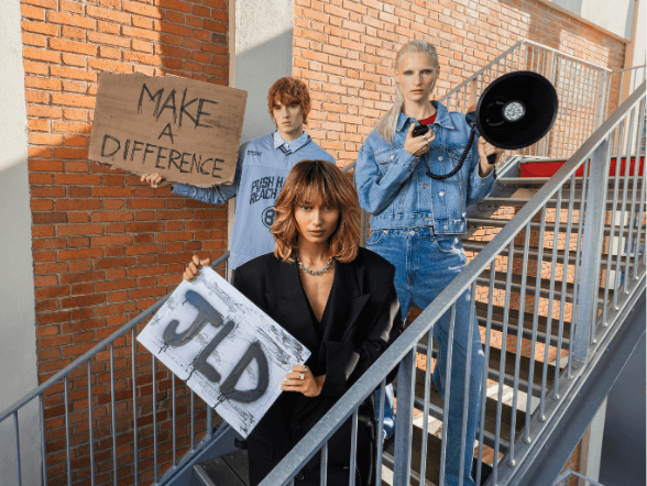 Trois mannequins posent sur un escalier extérieur en briques, tenant des pancartes « Make a Difference » et « JLD », illustrant la campagne automne-hiver Jean Louis David axée sur l’affirmation de soi et le style.