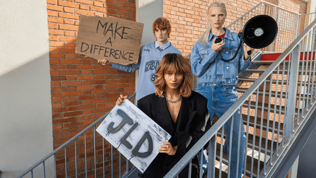 Trois mannequins posent sur un escalier extérieur en briques, tenant des pancartes « Make a Difference » et « JLD », illustrant la campagne automne-hiver Jean Louis David axée sur l’affirmation de soi et le style.
